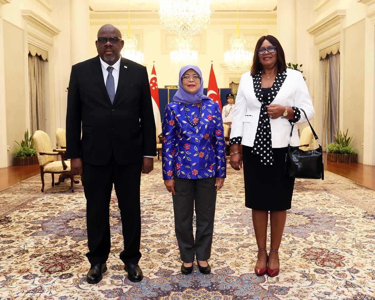 Three people stand in formal attire before Singapore and Turkish flags.